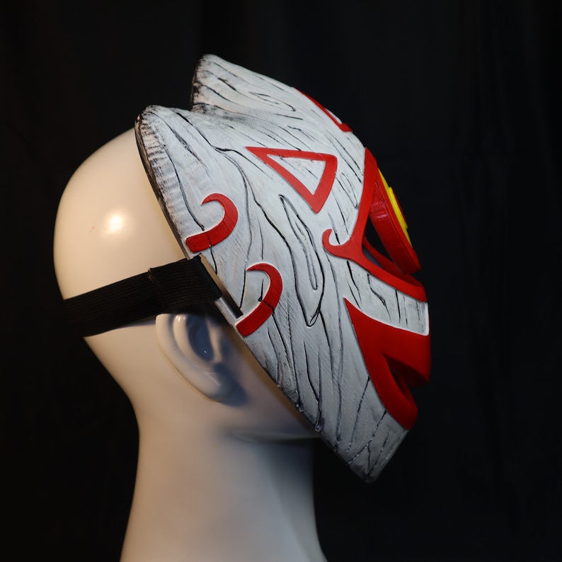 May include: A white and red mask with a wood grain texture and a red and yellow eye design. The mask has a black elastic strap.