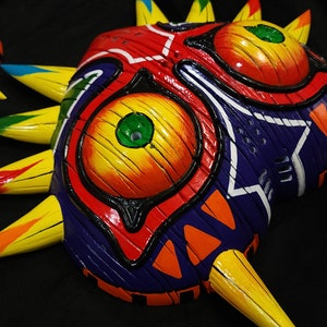 May include: A colorful, hand-painted mask with a sharp, pointed design. The mask features a red, yellow, and blue color scheme with green eyes and a black outline. The mask is made of wood and has a textured surface.