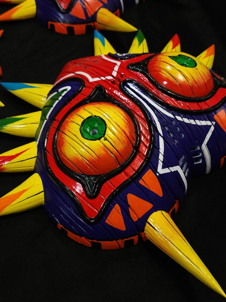 May include: A colorful, hand-painted mask with a sharp, pointed design. The mask features a red, yellow, and blue color scheme with green eyes and a black outline. The mask is made of wood and has a textured surface.