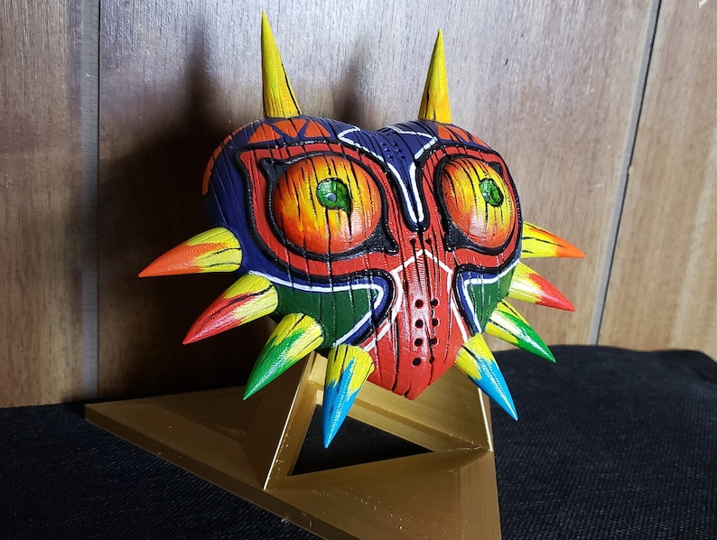 May include: A hand-painted, wooden Majora's Mask replica with a heart-shaped design. The mask is painted in vibrant colors, including red, orange, yellow, green, blue, and purple. It has a textured surface and sharp, pointed spikes around the edges. The mask is displayed on a gold-colored stand.