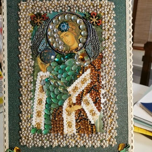 Peut inclure: Une œuvre d'art en perles vertes, brunes et blanches représentant une icône religieuse d'une femme avec des ailes. L'œuvre est encadrée d'une bordure en dentelle blanche et est exposée sur un chevalet en bois.