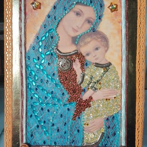 Puede incluir: Una obra de arte enmarcada que representa a una madre y un hijo. La madre lleva una túnica azul adornada con adornos turquesa y plateados. El niño lleva una túnica amarilla con adornos dorados y plateados. La obra de arte está enmarcada en un marco de madera marrón con un borde dorado.