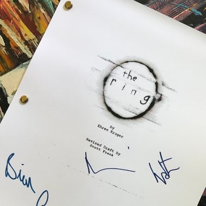 May include: A white shooting script for "The Ring" movie, featuring the title in a distressed circle design. The script is signed in blue ink and includes the date 10/3/01. The background is an abstract painting.
