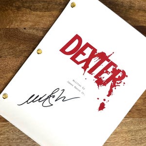 May include: A white script featuring the title "DEXTER" in red, with blood splatter details. The script is signed and includes the text "Written By James Manos Jr." It is held together with gold eyelets.