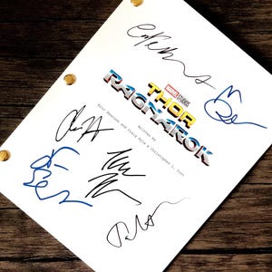 Può includere: Una sceneggiatura bianca con il titolo "Thor Ragnarok" e diverse firme in inchiostro blu e nero. La sceneggiatura è tenuta insieme da borchie dorate e poggia su una superficie di legno scuro.