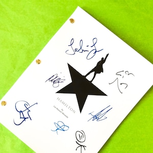 May include: A white paper with the title "HAMILTON" by Lin-Manuel Miranda, featuring a black star and silhouette, and multiple blue and black signatures. The paper is held together with gold brads and rests on a green surface.