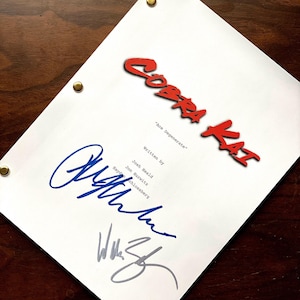May include: A white script titled "Cobra Kai" with red lettering and the subtitle "Ace Degenerate." The script is signed in blue and gray ink. The script is held together with brass fasteners and rests on a dark wood surface.