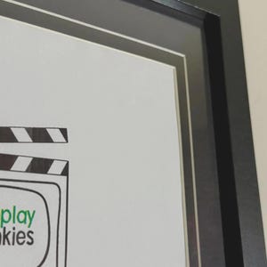 May include: A framed print with a black frame and white matting. The artwork features a cartoon television with the words "play" and "kies" in green text. A film clapperboard is above the television.