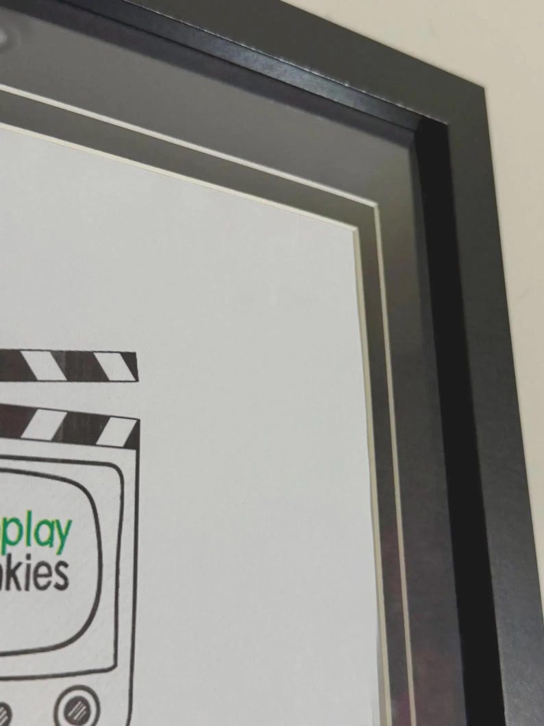 May include: A framed print with a black frame and white matting. The artwork features a cartoon television with the words "play" and "kies" in green text. A film clapperboard is above the television.