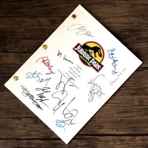 May include: A white script with the "Jurassic Park" logo and multiple signatures in various colors. The script is held together with brass fasteners and rests on a dark wood surface.