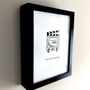 Op de afbeelding: Zwart ingelijste print met een witte achtergrond. De print toont een televisie met een klapbord erboven en de woorden "screenplay junkies" in een groen kader. Daaronder staat de tekst "Your script will go here."