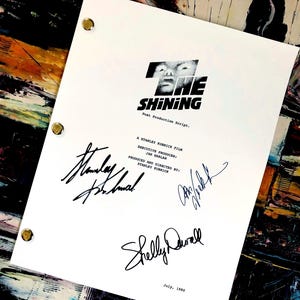 May include: A white "The Shining" post-production script with signatures. The title is in black, with a graphic of a face. The script is held together with brass fasteners. Signatures include Stanley Kubrick and Shelly Duvall. Dated July, 1980.