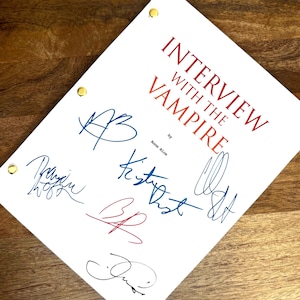 May include: A white script with the title "Interview with the Vampire" in red, and multiple signatures in blue, red, and black ink. The script is held together with gold brads and rests on a wooden surface.