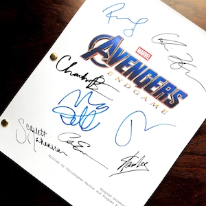 May include: A white script with the "Avengers: Endgame" logo and multiple signatures in black and blue ink. The script is secured with gold brads and rests on a dark wooden surface. Text at the bottom reads "Written by Christopher Markus and Stephen McFeely."
