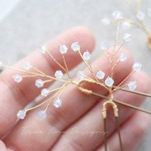 Op de afbeelding: Twee gouden haarspelden met delicate draadtakken versierd met kleine, heldere, iriserende kralen. De haarspelden worden in een hand gehouden.