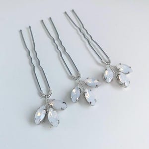 Op de afbeelding: Drie zilverkleurige haarspelden, elk versierd met een cluster van opalescente, ovaalvormige kristallen. De kristallen zijn gerangschikt in een delicaat, bladachtig patroon, wat een vleugje elegantie toevoegt. De achtergrond is wit.