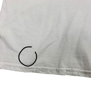 May include: Close-up of a white t-shirt. The fabric is slightly wrinkled, and the hem is visible at the bottom. There is a small stain near the bottom hem. The shirt appears to be made of a soft, lightweight material.