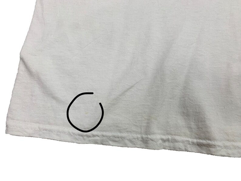May include: Close-up of a white t-shirt. The fabric is slightly wrinkled, and the hem is visible at the bottom. There is a small stain near the bottom hem. The shirt appears to be made of a soft, lightweight material.