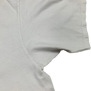 May include: Close-up of a white t-shirt, showing the shoulder and sleeve. The fabric appears to be a soft cotton, with visible seams and stitching along the sleeve edge. The shirt is plain and unadorned.
