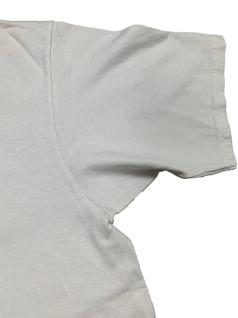 May include: Close-up of a white t-shirt, showing the shoulder and sleeve. The fabric appears to be a soft cotton, with visible seams and stitching along the sleeve edge. The shirt is plain and unadorned.