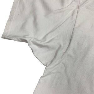 May include: Close-up of a white t-shirt sleeve. The fabric is slightly wrinkled, and the sleeve's edge is visible. The shirt appears to be made of a soft, lightweight material. The image is set against a plain white background.