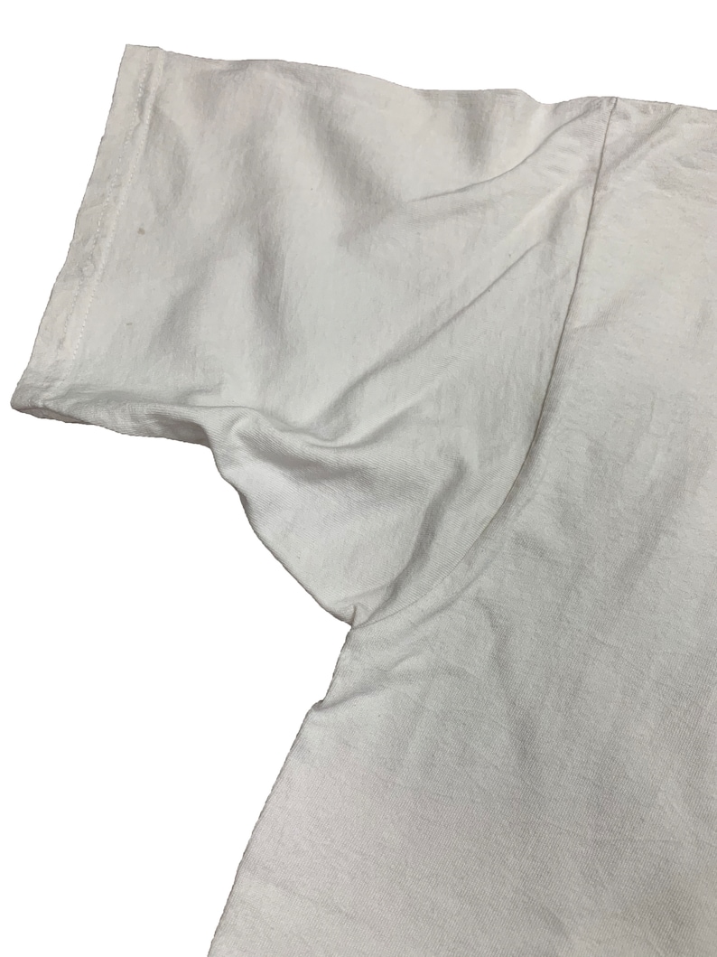 May include: Close-up of a white t-shirt sleeve. The fabric is slightly wrinkled, and the sleeve's edge is visible. The shirt appears to be made of a soft, lightweight material. The image is set against a plain white background.