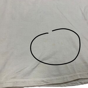 May include: A white t-shirt with a stain in the center. The fabric is slightly wrinkled, and the bottom hem is visible. The image is a close-up of the shirt's fabric.