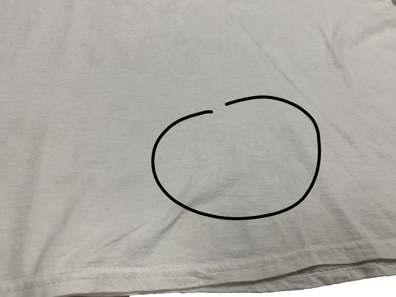 May include: A white t-shirt with a stain in the center. The fabric is slightly wrinkled, and the bottom hem is visible. The image is a close-up of the shirt's fabric.