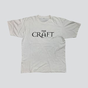 May include: White t-shirt with the text "THE CRAFT" in black, centered on the chest. Below the main text is smaller text. The shirt has short sleeves and a crew neck.