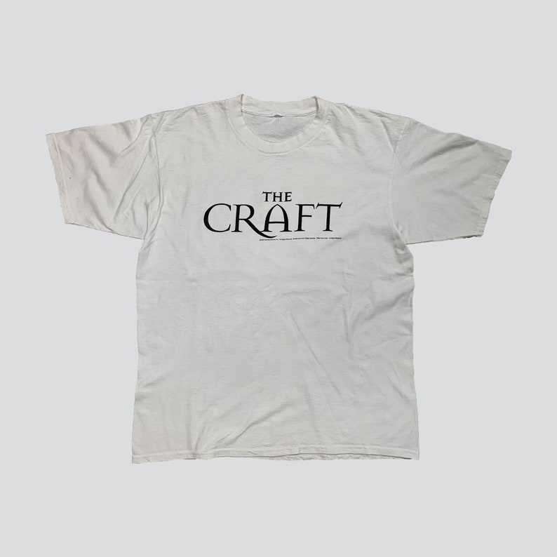 May include: White t-shirt with the text "THE CRAFT" in black, centered on the chest. Below the main text is smaller text. The shirt has short sleeves and a crew neck.