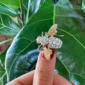 May include: A gold bee brooch featuring textured wings and a body encrusted with numerous small, clear, sparkling stones. The insect-shaped pin displays detailed metalwork on the wings and a dense cluster of stones forming the thorax and abdomen.