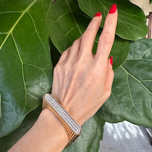 May include: A gold-toned bracelet with a rectangular face, adorned with multiple rows of sparkling, clear gemstones. The bracelet has a ribbed design on the sides. The background features large, green leaves.