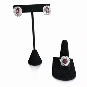 May include: A silver-toned jewelry set featuring a ring and matching earrings. Each piece has a central, square-cut, deep pink gemstone surrounded by a halo of small, clear crystals. The earrings are studs, and the ring is displayed on a black stand.