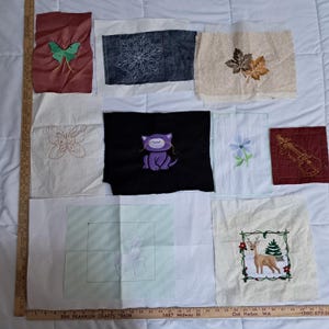 May include: A collection of nine embroidered fabric squares with various designs, including a butterfly, a cat, a flower, a violin, a deer, and a leaf. The squares are arranged in a grid pattern on a white surface.