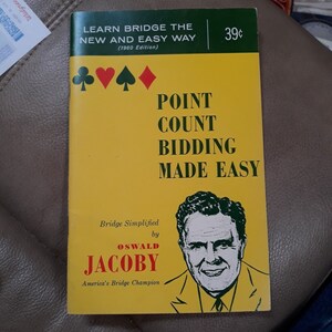 May include: A yellow book titled "Point Count Bidding Made Easy" by Oswald Jacoby, America's Bridge Champion. The book cover features a portrait of Jacoby and the four card suits: clubs, hearts, spades, and diamonds.