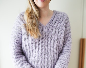 May include: A soft, lavender-colored crocheted sweater with a V-neck and long sleeves. The sweater is made with a textured stitch and has a relaxed fit.