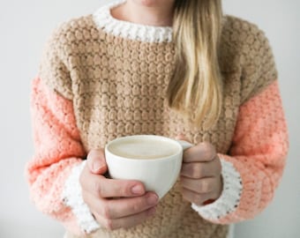 Puede incluir: Una mujer lleva un suéter de punto beige, rosa y blanco y sostiene una taza blanca llena de una bebida marrón claro.