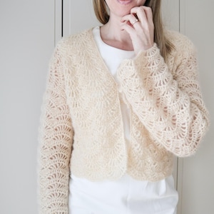 Pode incluir: Um casaco de malha de cor creme com um padrão de renda delicado. O casaco tem um decote em V e mangas compridas.