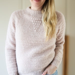 Puede incluir: Un suéter de punto de color rosa claro con un diseño texturizado en la parte delantera. El suéter tiene un cuello alto y mangas largas.