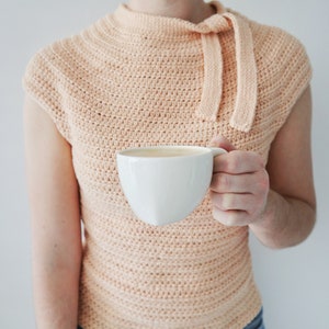 May include: A peach-colored, sleeveless crochet top with a mock neck and bow detail. The top is paired with blue jeans. A white coffee cup is held in the hand.