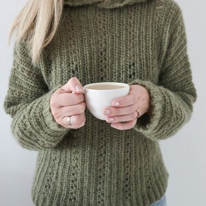 May include: A woman wearing a green knitted jumper with a hood, holding a white ceramic mug.