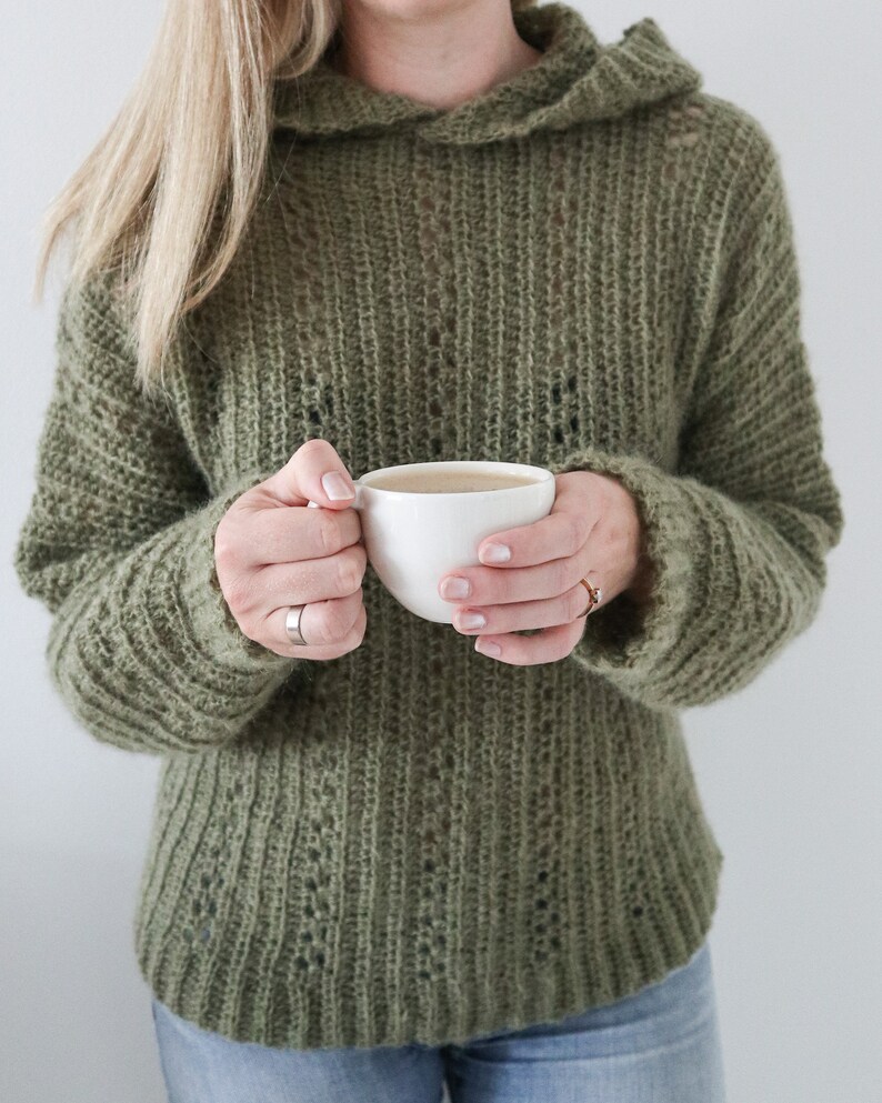 May include: A woman wearing a green knitted jumper with a hood, holding a white ceramic mug.
