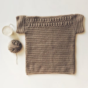 Peut inclure: Un haut crochet&eacute; marron &agrave; manches courtes avec un motif de point d&eacute;coratif autour de l'encolure. Une pelote de laine marron et une tasse de caf&eacute; sont &eacute;galement pr&eacute;sents sur l'image.