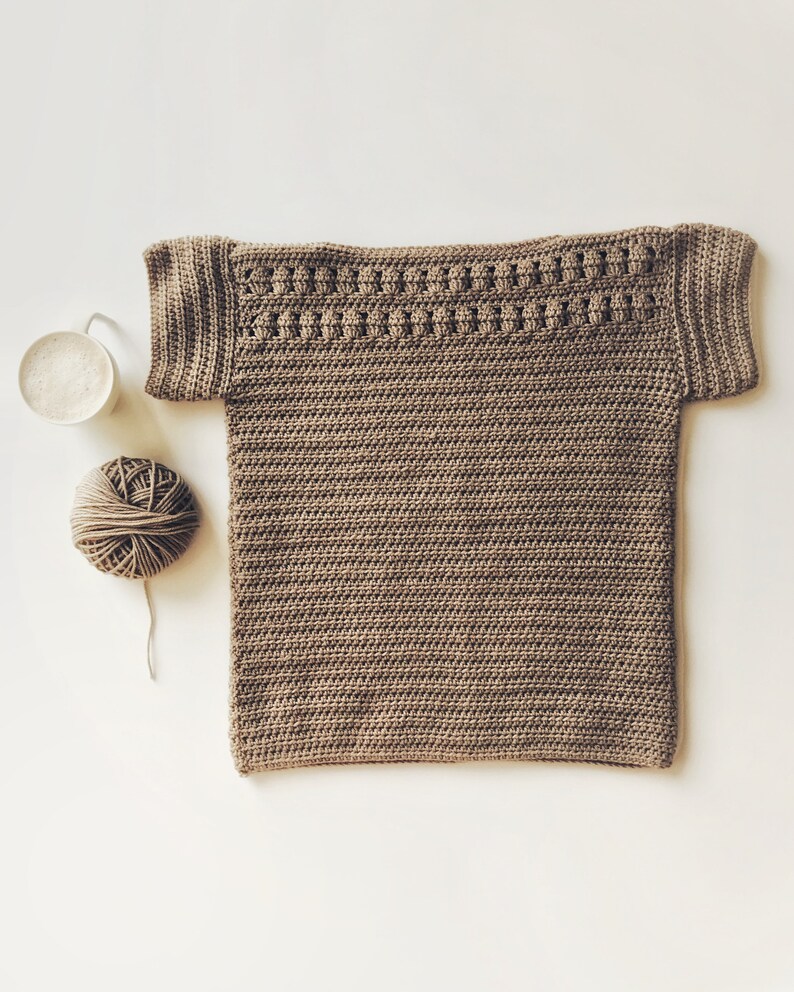 Peut inclure: Un haut crochet&eacute; marron &agrave; manches courtes avec un motif de point d&eacute;coratif autour de l'encolure. Une pelote de laine marron et une tasse de caf&eacute; sont &eacute;galement pr&eacute;sents sur l'image.