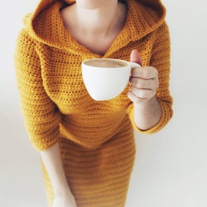 May include: A woman wearing a yellow crocheted hooded dress holds a white mug of coffee.