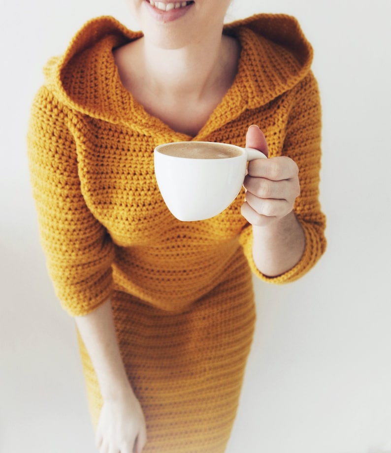May include: A woman wearing a yellow crocheted hooded dress holds a white mug of coffee.