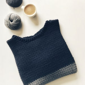 May include: A dark blue crocheted sweater with a gray crocheted trim. The sweater is laying flat on a white surface. There are two balls of yarn and a cup of coffee in the background.