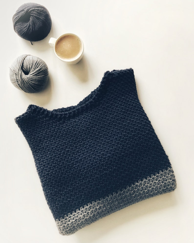 May include: A dark blue crocheted sweater with a gray crocheted trim. The sweater is laying flat on a white surface. There are two balls of yarn and a cup of coffee in the background.