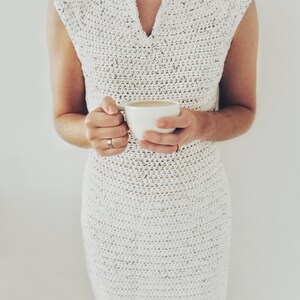 May include: A white crochet dress with a v-neck and sleeveless design. The dress is crocheted in a fine stitch and has a loose fit. The model is holding a white mug in her hands.