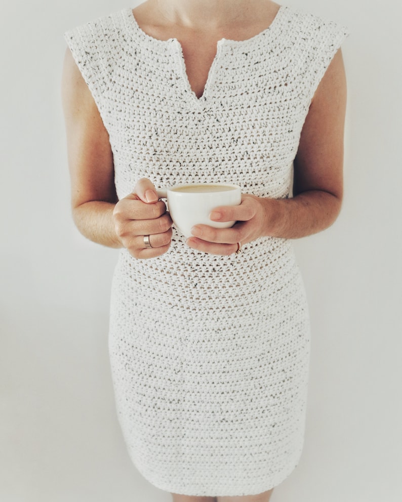 May include: A white crochet dress with a v-neck and sleeveless design. The dress is crocheted in a fine stitch and has a loose fit. The model is holding a white mug in her hands.